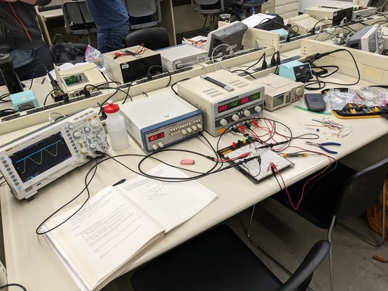 An electronics lab bench with an open lab book, circuit on a breadboard, tools, and parts. There is also an oscilloscope, signal generator, DC power supply, multimeter, and soldering station.