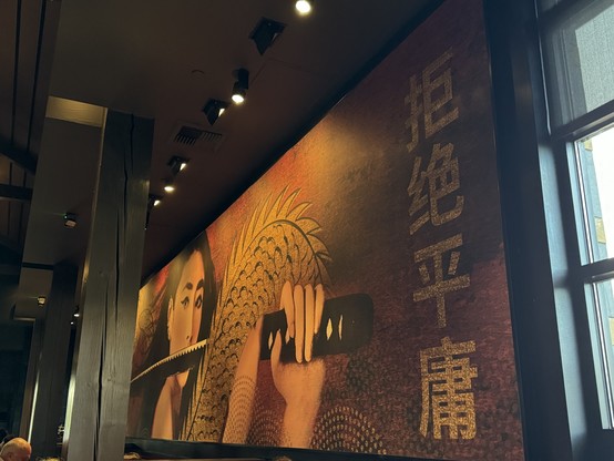 Interior of a PF Chang's restaurant with a large Asian art mural on the wall featuring an illustrated woman, decorative patterns, and Eastern Asian characters. There's a window to the right, and ceiling lights above.