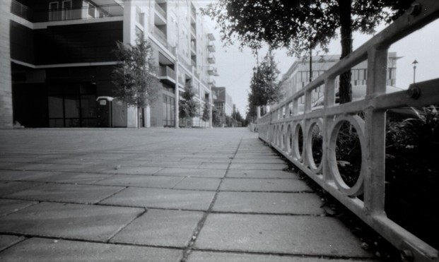 Black and white photograph of a brick walkway between planters and buildings.
