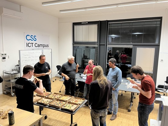 Some of the attendees standing around a table full of pizza, talking and eating.