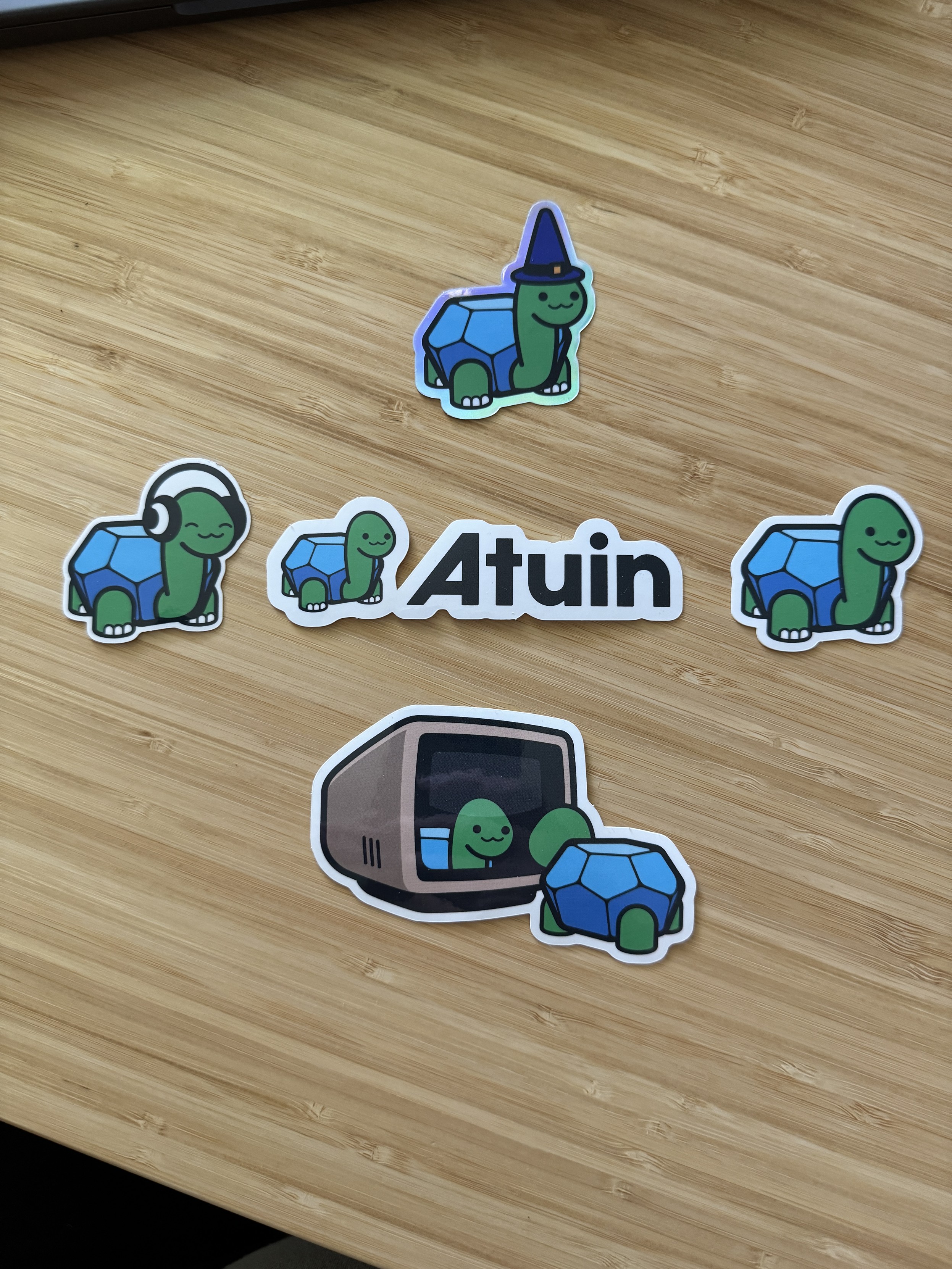 Five stickers from Atuin (Sync, search, and backup shell history with Atuin) on a wooden desk. All stickers have a cartoon turtle. One is wearing a wizard hat, another headphones, and one is looking at its reflection in a computer monitor. 