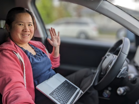 Mariatta, wearing pink hoodie and blue shirt, seated inside a car with laptop and steering wheel in front of her.