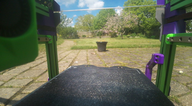View from inside the housing of a Voron 0.2 3D printer. In the background is an apple tree in blossom on a lawn. The sky is blue with white fluffy clouds. The printer is sat on a stone paved patio.