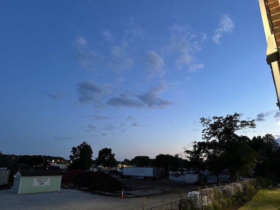 Twilight sky over a construction site with clouds, a fenced area with trailers, and a large tree on the right.