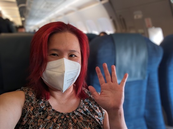 Mariatta selfie seated inside the plane, waving. Red haired woman with a white kn95 mask