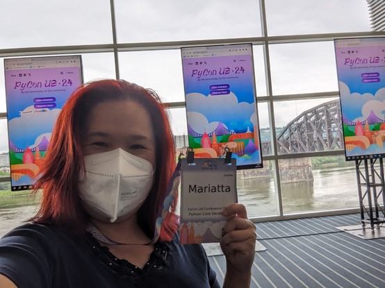 Selfie of Mariatta, red haired woman with KN95 mask holding the PyCon US conference badge.
Behind her are 3 monitors with PyCon US 24 landing page displayed