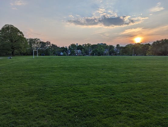 A view of a soccer field, with houses, trees, and the setting sun. Behind the trees, in the distance, is a city skyline.