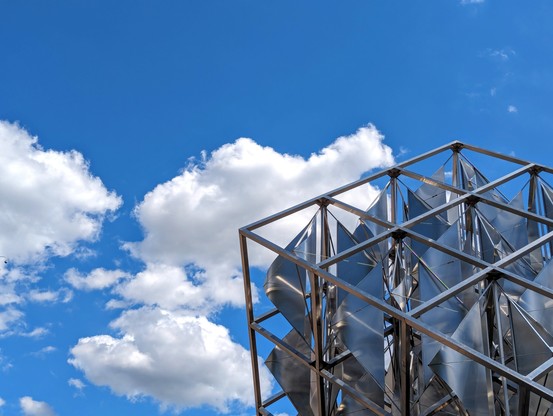 A metal structure formed of square grids with curved metal foils that spin in the wind gleams in the sunlight set against a bright blue sky with fluffy white clouds. 