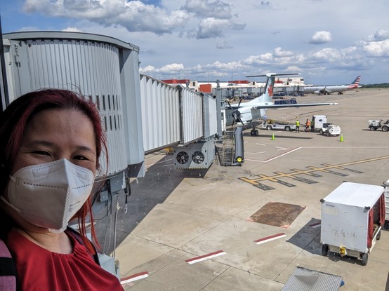 Selfie of Mariatta in front of airport gate with a plane behind her.
Red haired woman wearing a white kn95 mask