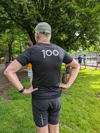 A post-run photo of the back of a mid fifties guy in a black parkrun 100 tee shirt. His hands are on his hips. He's also wearing a beige cap and black trail shorts. It's a sunny day. The trees are casting shade onto the grass. Various other runners and volunteers are in the background.
