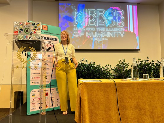 Woman standing on stage in front of a slide reading "Mirror, mirror: LLMs and the illusion of humanity".