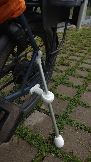 Back wheel of an e-bike. Instead of a typical kick-stand, a mostly 3d-printed contraption is attached. Where the kickstand would normally be fixed (side of the frame), a 3d printed threaded rod is attached with a ball joint, where an aluminum rod is passed through. The top of the aluminum rod loosely clamps to a part of the bike frame a bit higher up with a 3d printed u-shaped part, the bottom of the rod has a teardrop-shaped 3d printed bumper slid on and balances the bike so it doesn't fall. T…