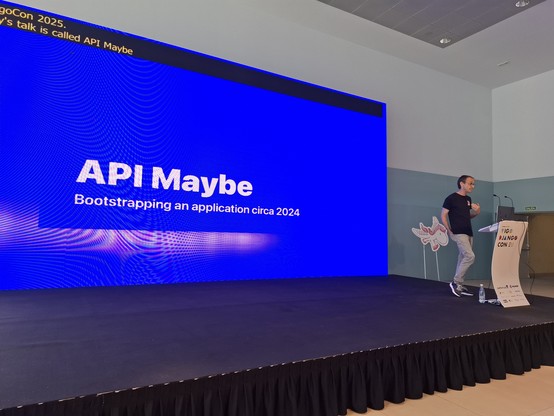 Carlton Gibson on the Django Con Europe stage with blue slide in the background saying "API Maybe: Bootstrapping an application circa 2024".