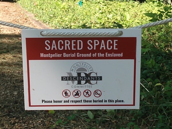 A rectangular sign suspended from a rope across a trail. The sign says:

SACRED SPACE
Montpelier Burial Ground of the Enslaved

MONTPELIER
DESCENDANTS
COMMITTEE
[superimposed on large letters 