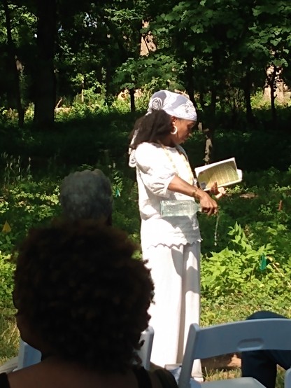 A woman dressed in white (Rashida Kirton, of the Montpelier Descendants Committee) stands outdoors in bright sunlight in front of rows of people seated in chairs. Behind her we see a green wooded area (part of the burial ground). In one hand the woman holds an open book. In the other hand she holds a clear glass bottle full of water. She is pouring water from the bottle onto the ground.