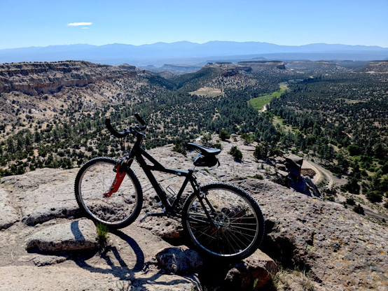 A mountain bike on the edge of a mesa. Below you can see the confluence of two canyons, with a grassy streambed in one, and another finger mesa coming in from the left. Hazy mountains (the Sangre de Cristos) in the distance.