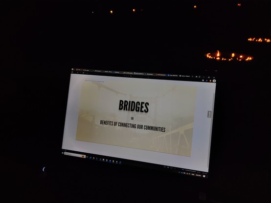 Foreground is a laptop with a presentation slide that reads "Bridges, or Benefits of Connecting Our Communities. The rest is dark, with exception to the firepit seen in the background.