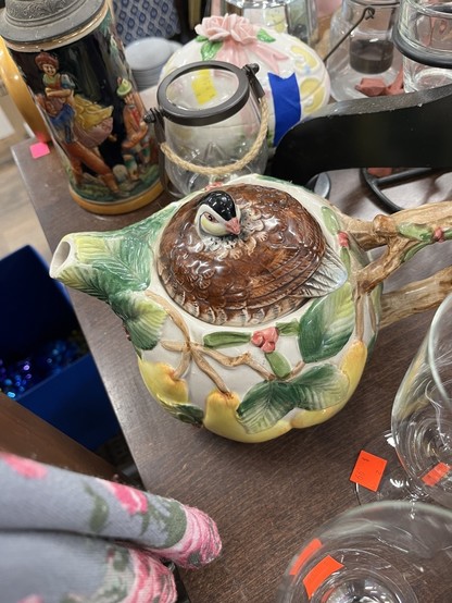 A ceramic teapot shaped like a perched bird amidst various household items on a table at a thrift shop. 
