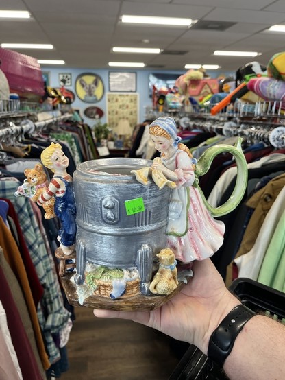 A hand holding a decorative teapot with figurines, of a bow holding a teddy bear as the spout and a woman wearing a dress with its ribbons as the handle in a thrift store.