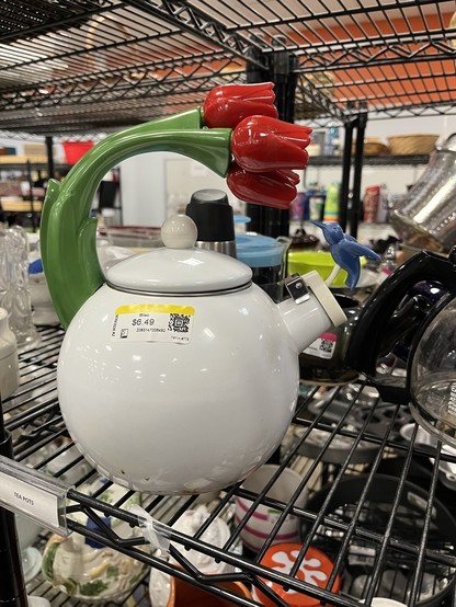 A white teapot with a decorative tulip-shaped handle and a small hummingbird figure sits on a thrift store shelf. 