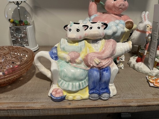 Ceramic cow figurines knitting together on a shelf among various decorative items, including a pig in a bath tub, gum ball machine and a pink glass basket.