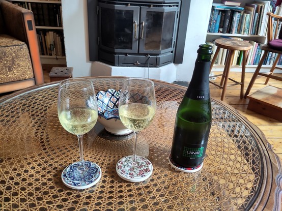 Photo. Two large wine-glasses filled with bubbly yellow wine stand on ceramic coasters with Turkish floral patterns. On the third coaster, a green bottle of Portuguese cava, L'ANAE brand. The round table is of wicker covered with a glass plate. In the background, a patterned bowl containing the rest of the coasters. Behind that, a wooden chair and a fileplace surrounded by bookshelves.
