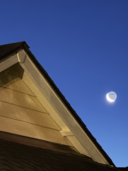 A 4 a.m. photo of the rising crescent moon. The dark side of the moon with its multiple craters are somewhat visible against a dark blue backdrop of morning sky. A yellow house's rooftop is in the foreground.