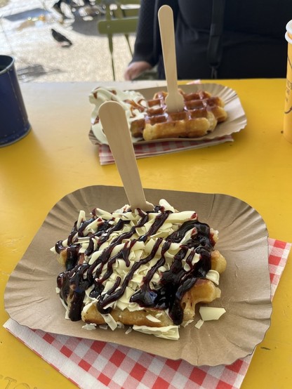Two waffles on a yellow table, one topped with white cream and chocolate, the other with caramel, accompanied by wooden utensils.