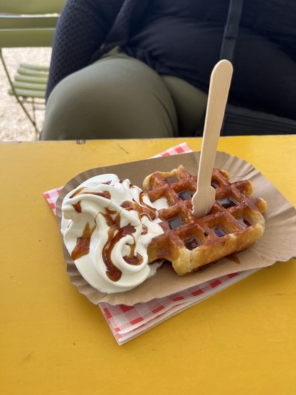 A waffle topped with whipped cream and syrup, with a wooden spoon, rests on a yellow table, suggesting a casual outdoor eating setting.