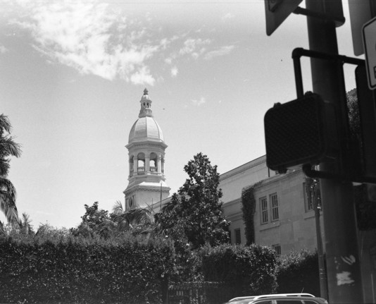 A black and white photo of a building against a clear sky with sparse clouds. In the foreground there is a pedestrian traffic light and some bushes surrounding a small gated park 