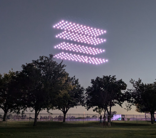 The end of a drone show, in twilight, with the drones returning to their landing pads, visible as four layers of 5 x 13 white lights. In the foreground are trees and some spectators.