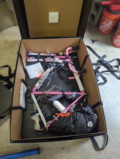 A pink bicycle, partly disassembled, in a large box.