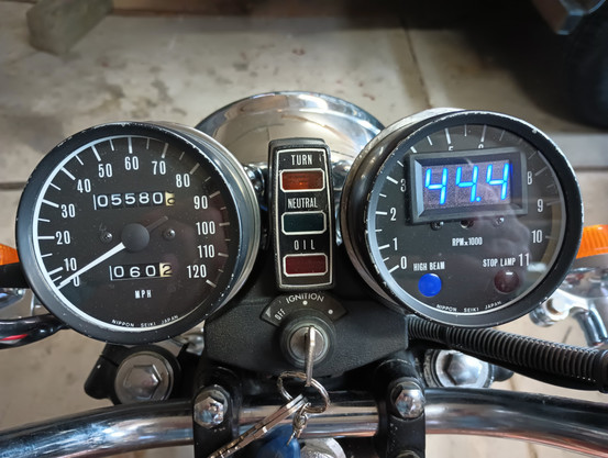 Color photo of the dash panel on an old, 1970s era motorcycle. On the left is a round white on black speedometer indicating 0mph, 5580.9 total miles, and 60.2 trip miles. On the right is a round white on black tachometer, except the tachometer needle has been removed and a rectangular volt meter has been fitted inside the housing under the glass cover. The meter is on indicating 44.4 in blue digital numbers. A high beam light and stop lamp are at the bottom on the gauge and both lights are off. In the middle is a rectangular light bar with a yellow turn light at the top, green neutral light below that, and red oil light below that. All three lights are off. A key switch is in the middle showing 'off' and 'ignition' and an unlabeled third dot on the right. Parts of the two turn indicator lights are seen on the left and right. The top of the chrome headlight is at the top of the frame and a small portion of the handlebars is at the bottom.