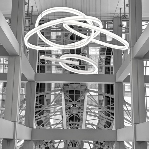 Black and white photo of lighted ring structures suspended form a high ceiling framed by concrete pillars and beams behind which there's a metal archway lattice which is partially visible. 