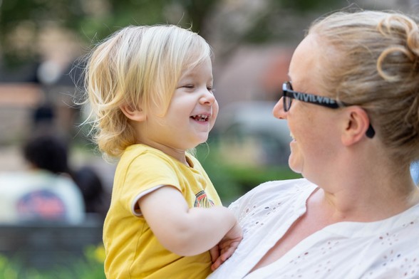 A smiling white woman (Kirstie) wearing glasses holds a laughing toddler (her daughter) with blonde hair and a yellow shirt in an outdoor setting.