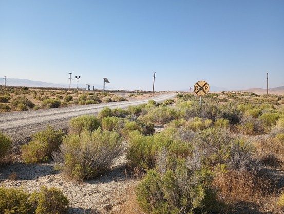 A color photo of a railroad crossing in a desolate landscape. There's a round yellow R & R crossing sign and a small red stop sign in the distance. Along the distant tracks running from left to right are a couple of old telegraph poles and a solar powered track light.