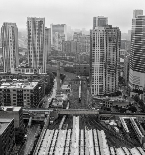 A black and white top down view of a train station from a high vantage point showing a train heading in through one of many winding tracks surrounded by tall buildings.