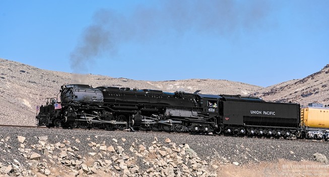 A color photo of a very large black steam locomotive. Some visible smoke is coming from the smoke stack. Behind the loco of a large black tender car and behind that is a bit of another yellow colored car. The loco is running across the frame from right to left. It is high on a embankment with gray rocks on the side. Rocky hills are visible in the near background.