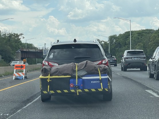 A vehicle driving down the highway.  To the back is mounted a pair of large boxes.  On top of the boxes is a roughly six foot long bag in the shape of a human body.

Wait... seriously.  Is that a human body?