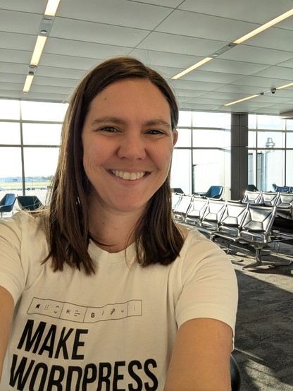 Amber wearing her Make WordPress Accessible shirt taking a selfie in front of a sunlit airport gate seating area.