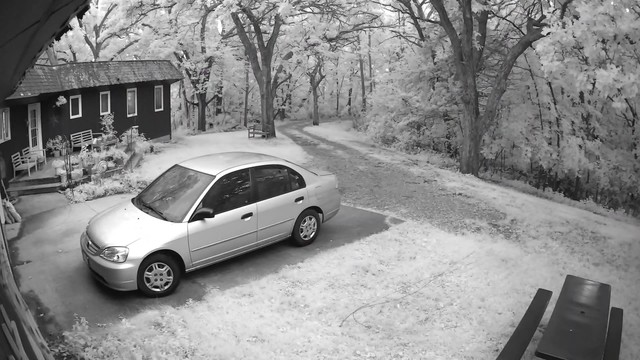 An infrared photograph of a house, car and driveway. The photo is black and white, and the trees that surround the house and driveway have foliage that is bright white against the darker trunks and branches. 