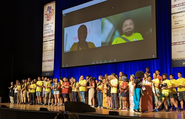 Many people in colourful t-shirts on a stage. Behind them are banners with the EuroPython 2024 logo and a screen showing two more people joined via a Zoom call.