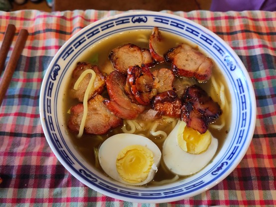 On a checkered table cloth, a blue/white chinese bowl filled with stock, char siu, eggs. The stock is darkish, the eggs are well-cooked (because runny eggs are icky).