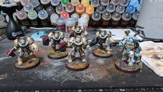 A squad of 5 Deathwing Terminators in their classic bone coloured armour, and a Dark Angels Apothecary Biologis. My preferred Dark Angels scheme tends towards Disciples of Caliban, so the left shoulder has a black rim (albeit with the standard DA Winged Sword because freehand rampant lions with swords is beyond me, and the 30k transfer sheet doesn't contain enough). They are standing on a well used painting mat, and behind them are arrayed a bank of colour ordered paints.