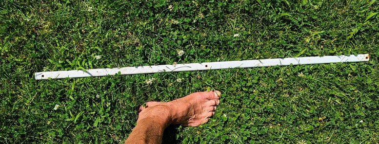 Section of straight white painted metal stake. It has plastic plugs at each end. The stake is rounded rectangular in cross section and a bit over a meter long. There is a foot in the image for scale.