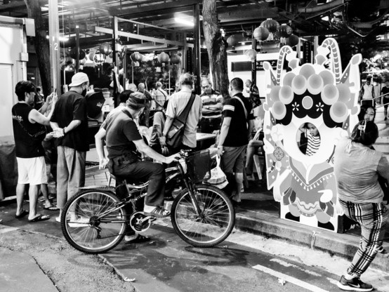 On the left cyclist and onlookers checking out a checkers match, on the right preparing to take photo behind a cardboard stand of a Chinese opera singer. 