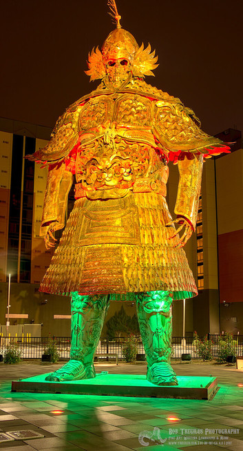A very tall metal sculpture of a skeleton in an ancient Asian war armor. It's night time photo and the lighting makes the top part down to the bottom of the 