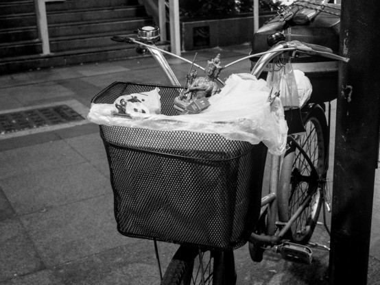Black and white photo of a parked bicycles with a small Buddha statue in the carrier basket and a bag of black coffee drink hangs from the left handle bar. 