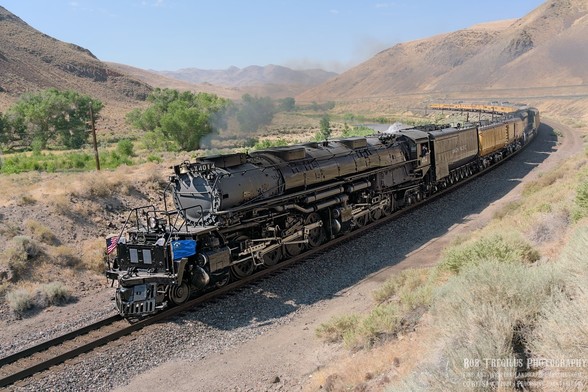 A color landscape photo of an old steam engine pulling about a dozen yellow train cars. The locomotive is turning to the left along a bend in the tracks. The landscape is high desert and dry brush. Low mountains are seen on both the right and left of the frame. There are a few trees in the background growing alongside a river that is barely visible on the opposite side of the train. A column of black smoke emanates for the locomotive under a blue sky.