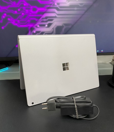 A grey laptop is positioned on a desk, facing away from the camera, featuring a Microsoft logo. In front of the laptop is a power adapter with a coiled cord. In the background, there is a monitor displaying a purple circuit-like pattern.